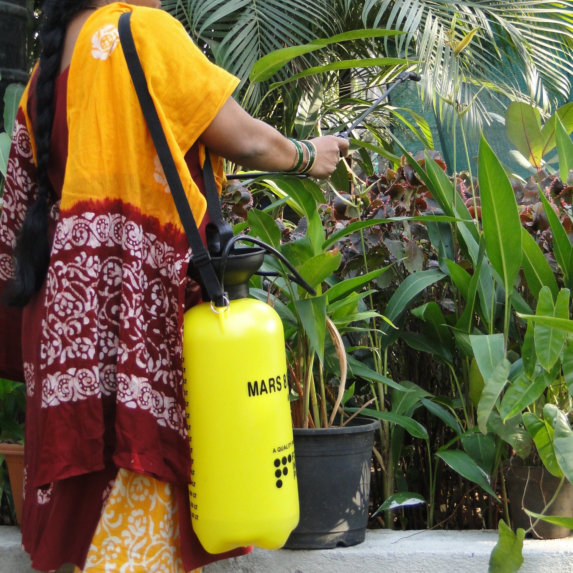 Woman in a yellow and red traditional outfit using a yellow MARS spraying can on plants.