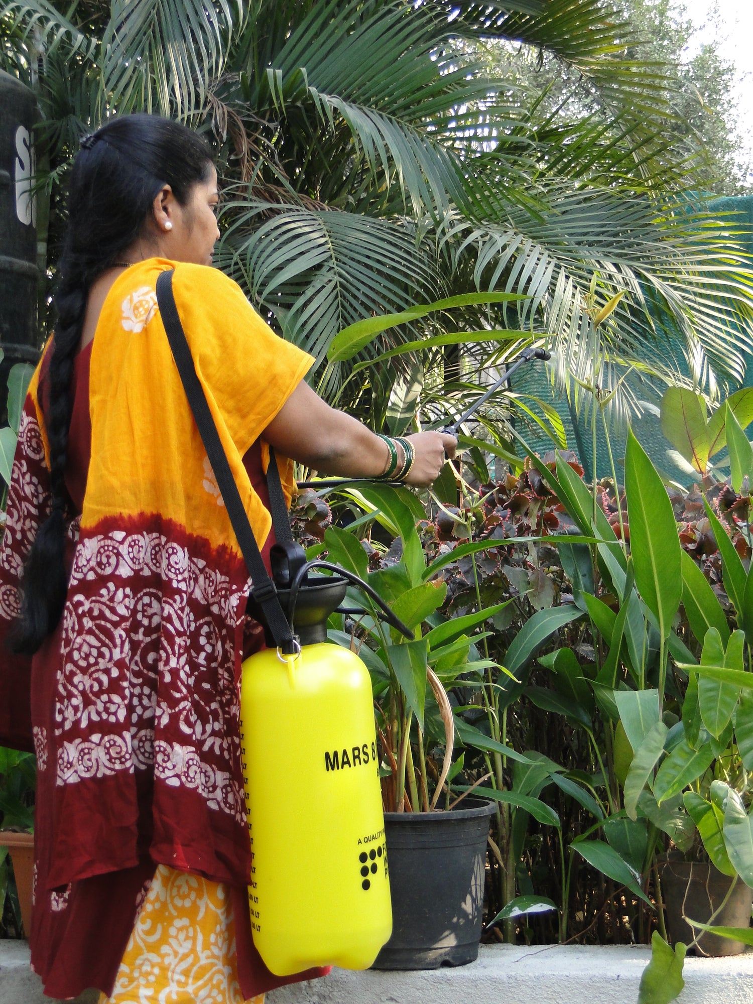 Woman in a yellow and red traditional outfit using a yellow MARS spraying can on plants.